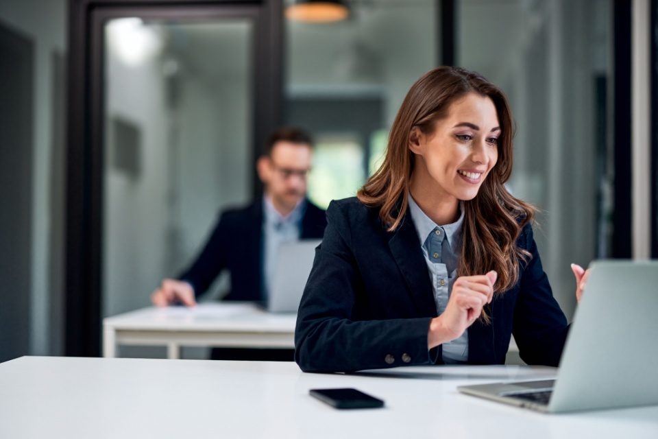 A businesswoman in a suit looks with delight at the laptop screen on her desk, while a male colleague works at his desk behind.